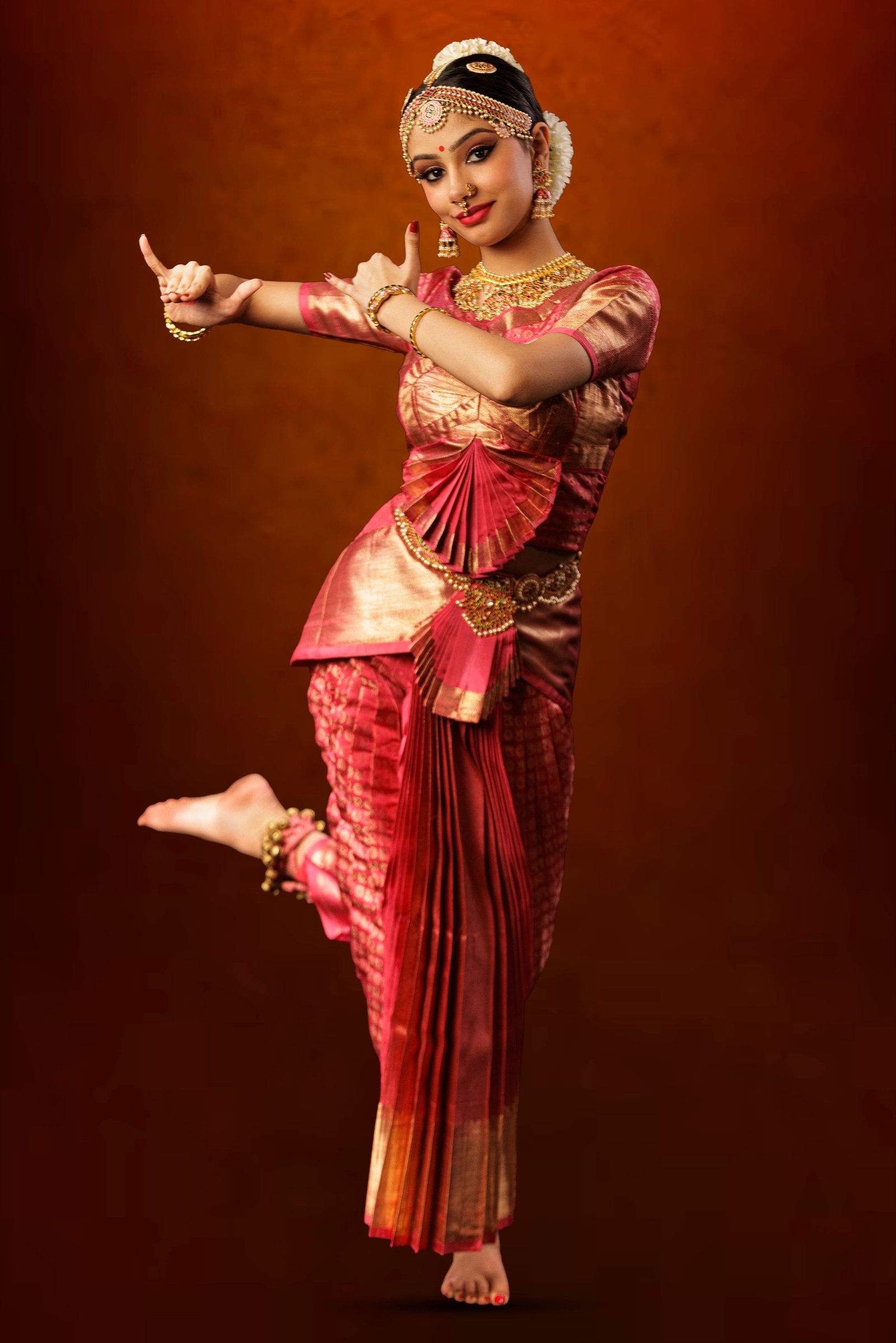 Ilesha Singh in rose-pink and gold silk Kuchipudi costume, one leg raised behind her, hands in raised gestures